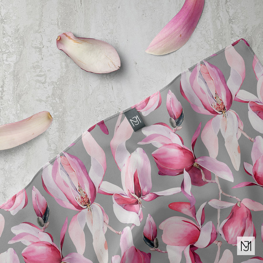 The Magnolia pattern on a draped silk scarf with a woven MJ label, next to loose magnolia petals on a stone surface.