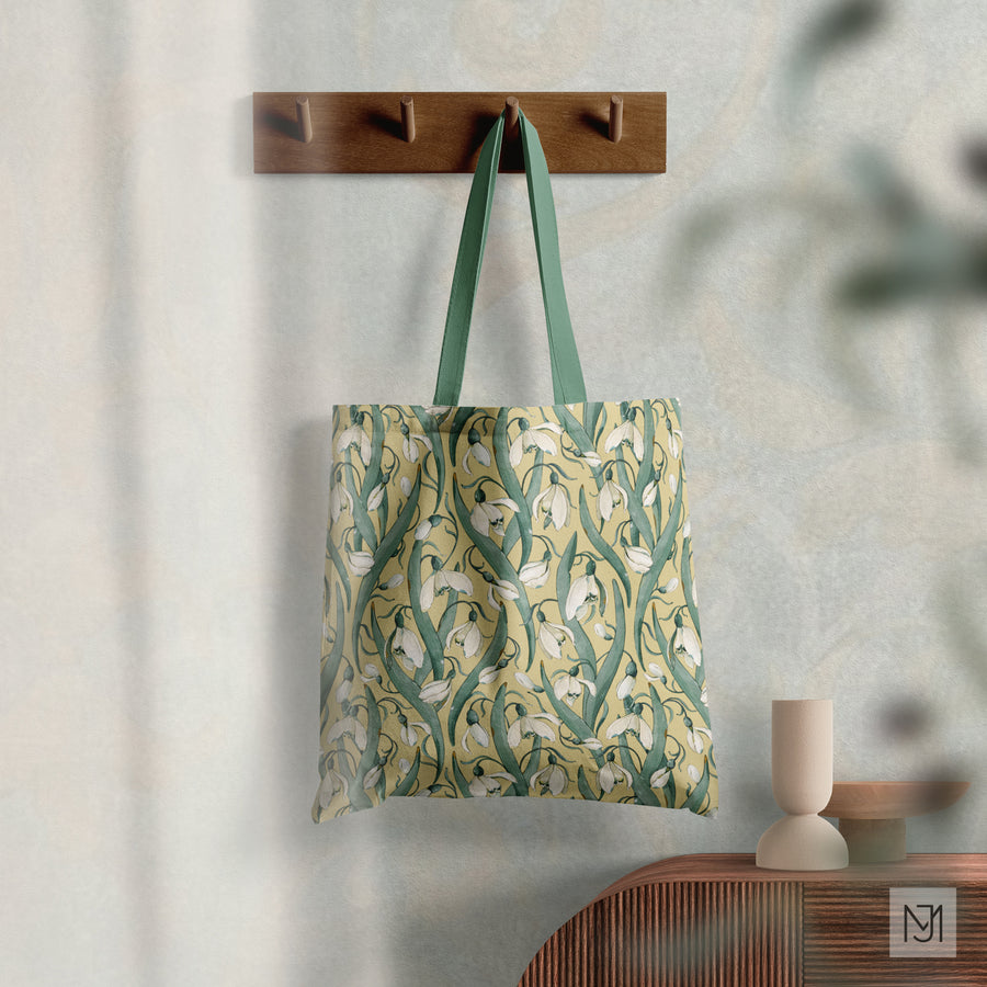 Tote bag by Media Jamshidi with a sage green strap hanging from a wall hook, in a pale yellow colorway of the snowdrop pattern