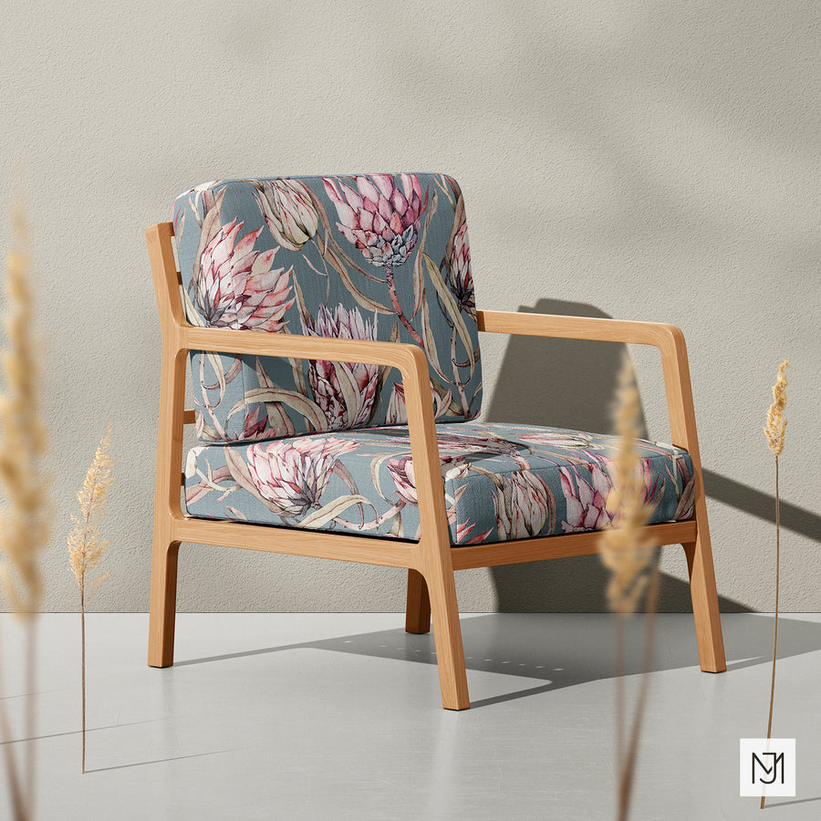 Tropical Protea pattern upholstered on a wood-framed armchair, shown against a beige wall with dried grass stems in the foreground.