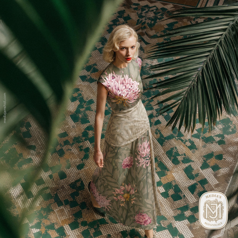 Woman wearing a sleeveless top and skirt set printed with the Quiet Garden pattern in a sage green colorway, standing on tiled floor among palm leaves