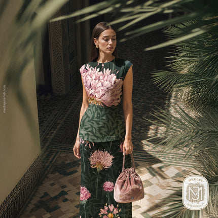 The same dark green cap sleeve top and skirt set in the Quiet Garden seamless surface pattern, worn in a different pose, standing among palm leaves in a tiled courtyard with a pink velvet bag in hand.