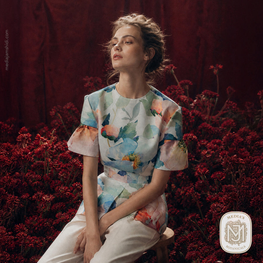 Woman wearing a puff sleeve blouse and pants in the Spring Waltz pattern, seated among dark red flowers