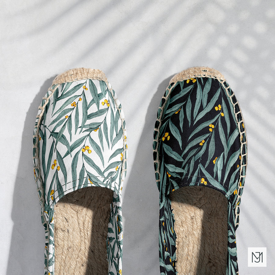Golden Wattle Leaves pattern on a pair of espadrilles, one in white and one in black, both with green wattle leaves and yellow blossoms.