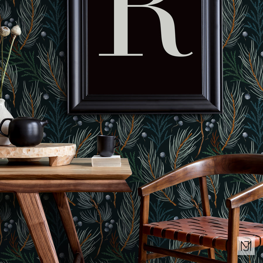Winter pattern as wallpaper behind a framed monogram print, a wood table with a black teapot and mug, and a woven leather chair.